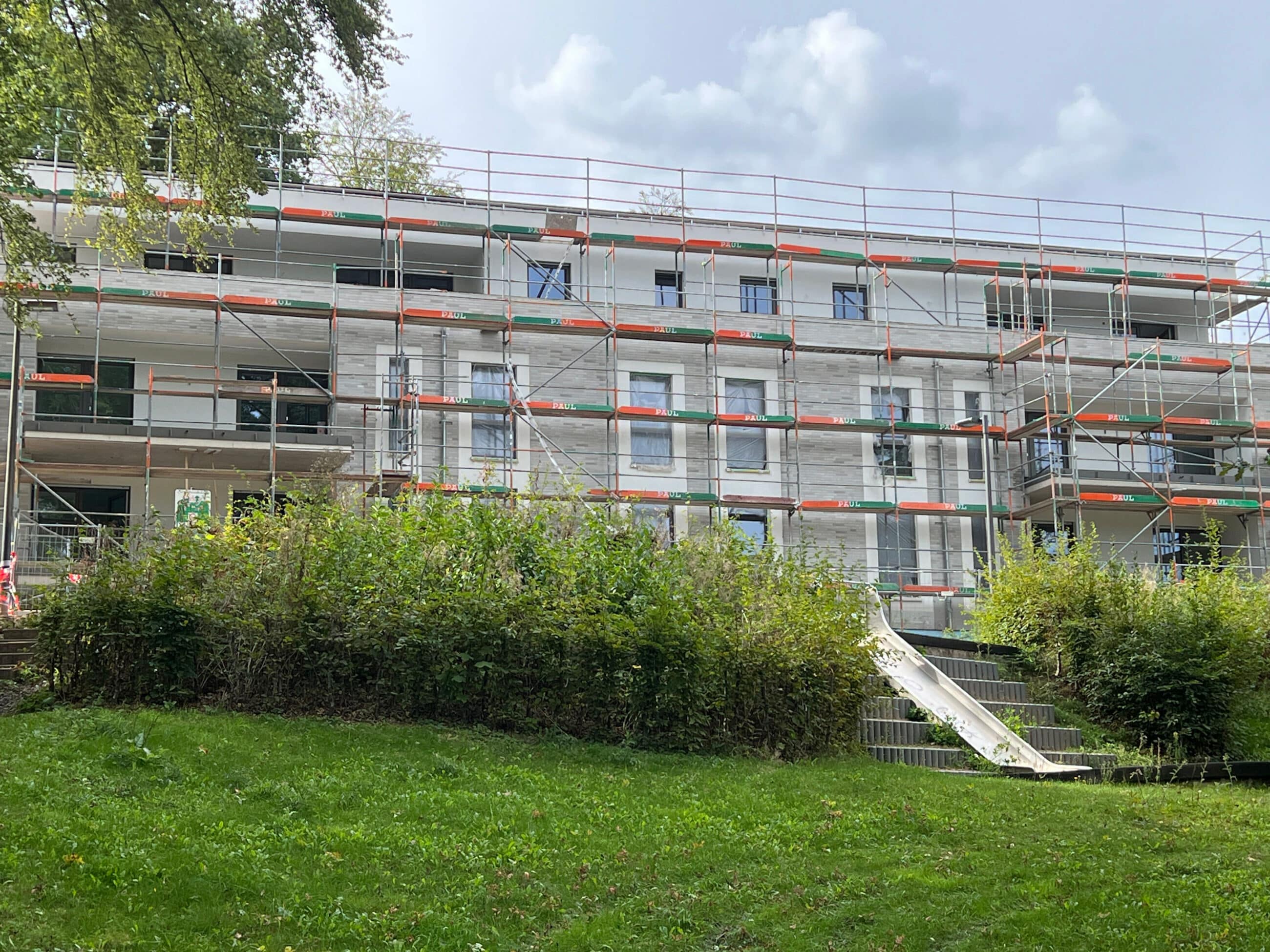 Ein im Bau befindliches dreistöckiges Gebäude, das mit Gerüsten umgeben ist, davor grüne Büsche und im Vordergrund eine Rasenfläche. Der Himmel ist bewölkt.
